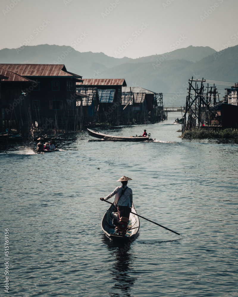 Inle Lake, Myanmar : December 9th 2019 : burmese woman wearing an asian conical hat paddling on a longboat on a canal to Inle Lake on a late afternoon with traditional houses on stilts.