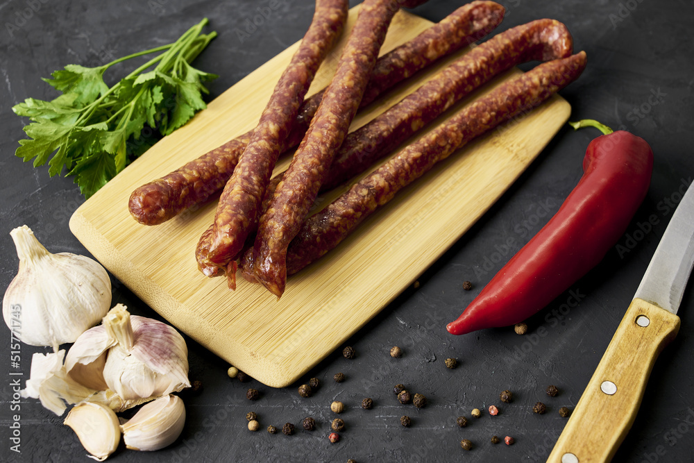 spicy smoked sausages on a cutting board next to red pepper garlic and herbs on a black background spicy smoked sausages on a cutting board next to red pepper garlic and herbs on a black background
