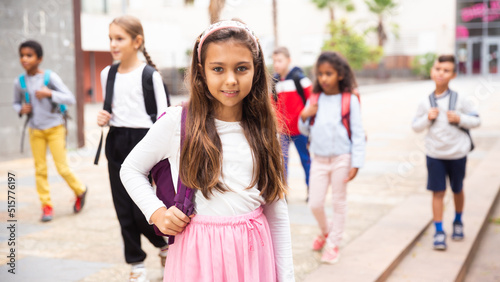 Portrait of tweenager girl with backpack walking with other schoolchildren to school campus after lessons on warm fall day.