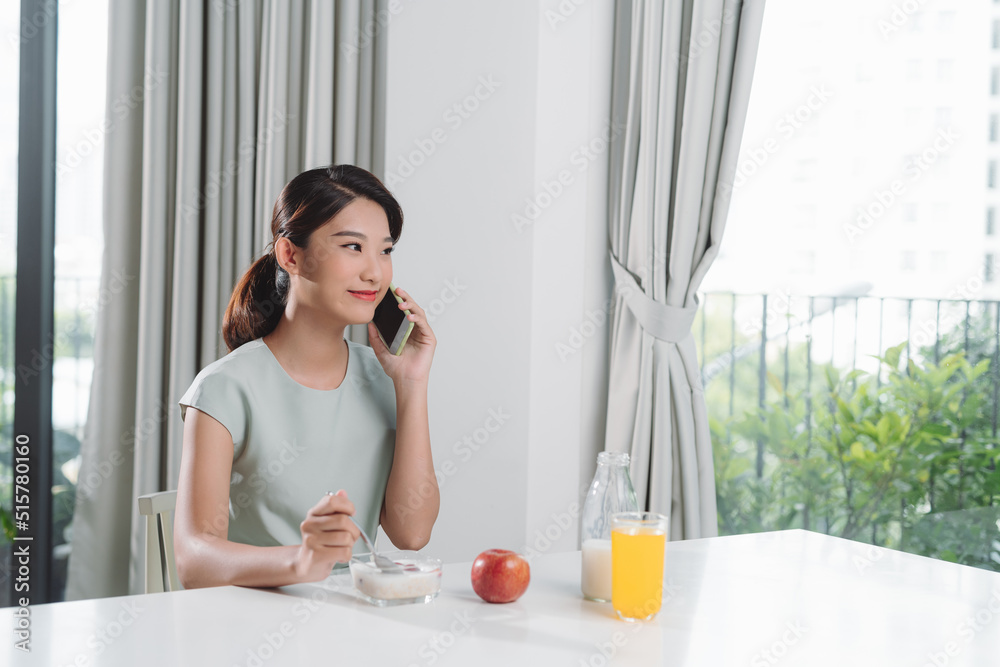 Beautiful woman eating tasty oat with milk while talking on mobile phone at table