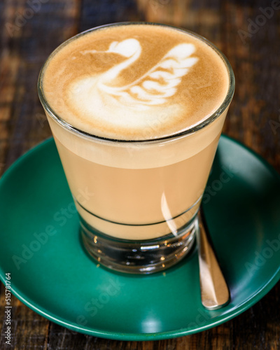 A latte foam art on a cup of coffee, placed in a drinking glass and green saucer with a wood grain background