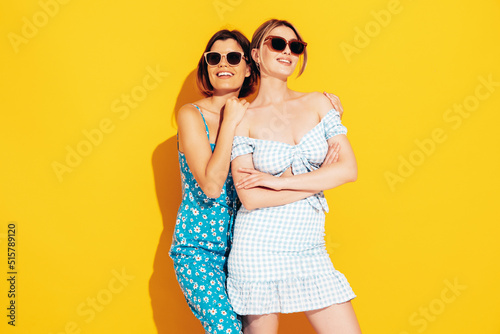 Two young beautiful smiling brunette hipster female in trendy summer dresses. Sexy carefree women posing near yellow wall. Positive models having fun. Cheerful and happy. Isolated