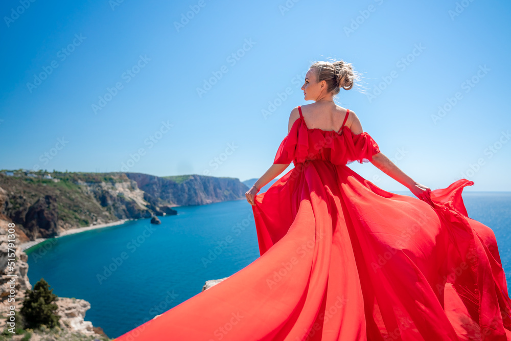Blonde with long hair on a sunny seashore in a red flowing dress, back view, silk fabric waving in the wind. Against the backdrop of the blue sky and mountains on the seashore. Blonde with long hair on a sunny seashore in a red flowing dress, back view, silk fabric waving in the wind. Against the backdrop of the blue sky and mountains on the seashore.