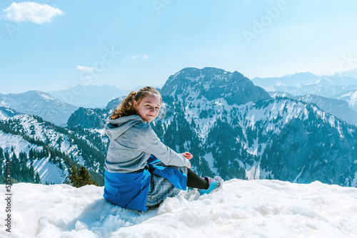 A young and joyful girl sits on the snow on top of a mountain and looks at the surrounding mountains.