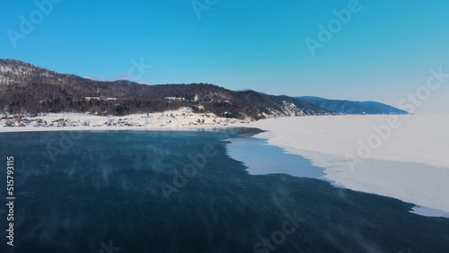 The river is split in half by ice. Moves towards the hilly bank. Along the lake there is a motorway with cars. The white snow contrasts with the dark blue water. Vapor rises over the icy calm surface