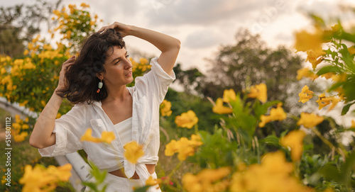 Stylish young caucasian woman adjusts her hair while looking to side among blooming flowers. Brunette wears white shirt in summer in nature. Weekend enjoyment concept