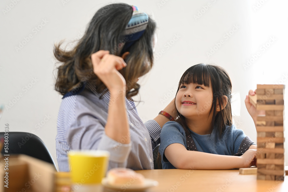 Little asian child and middle aged grandma enjoy playing wood blocks tower game in bright living room Little asian child and middle aged grandma enjoy playing wood blocks tower game in bright living room