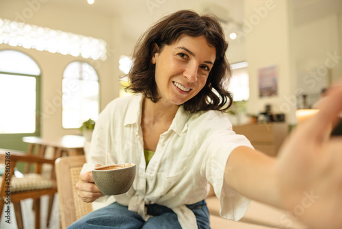 Happy young caucasian woman holding fragrant coffee and reaching for camera sitting indoors. Brunette wears casual clothes in spring. City life concept