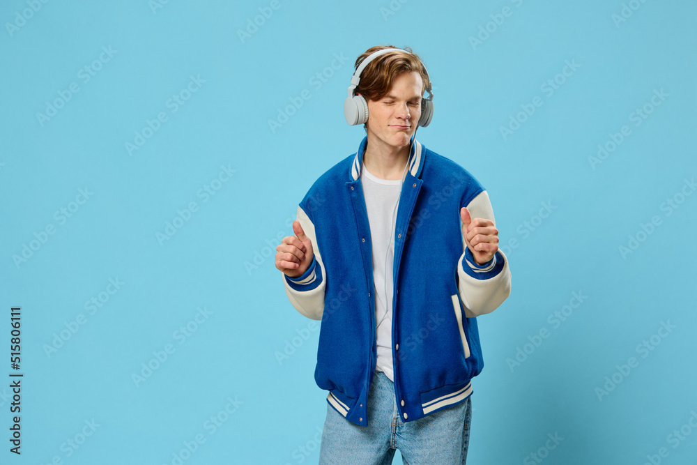 Fototapeta premium naughty handsome guy student stands dancing with hand movements on a light blue background listening to music through headphones