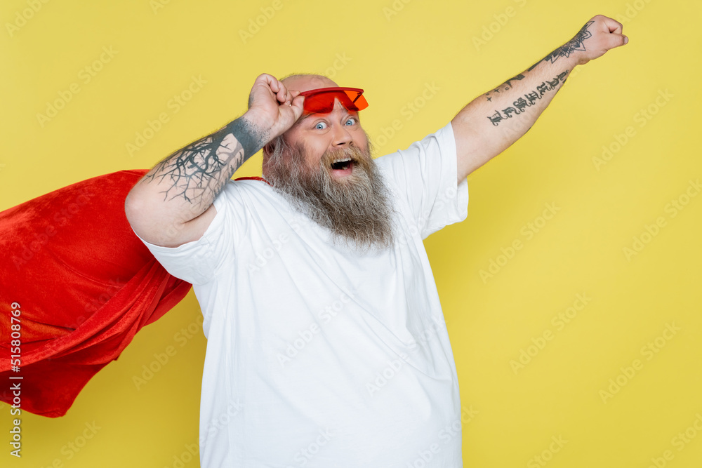 tattooed plus size man in superhero cloak posing with outstretched hand isolated on yellow. tattooed plus size man in superhero cloak posing with outstretched hand isolated on yellow.