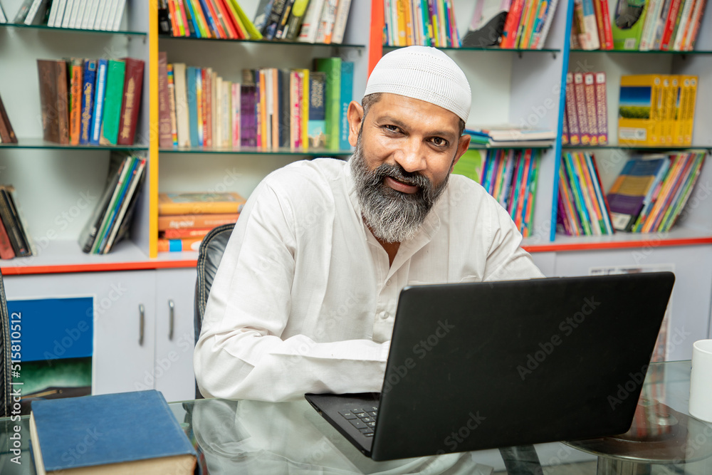 Portrait of happy asian muslim man working on laptop at desk in library with bookshelf behind him. browsing internet, studying online.