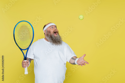 Photo amazed overweight man with racquet playing with tennis ball isolated on yellow