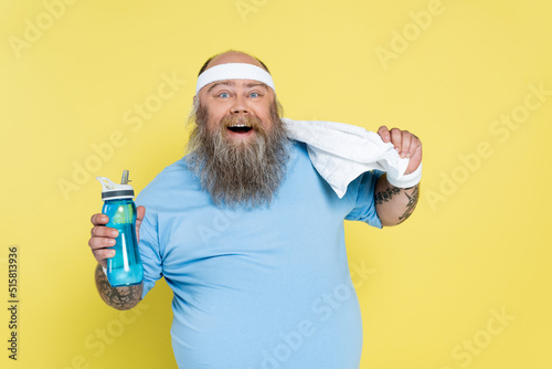 cheerful plus size man with sports bottle and towel looking at camera isolated on yellow.