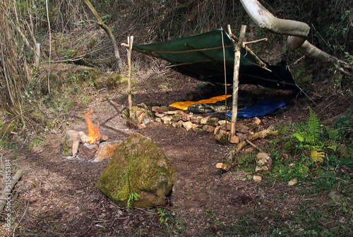 Bushcraft shelter built in the middle of an atlantic tree forest. Wooden shelter with a tarp and a fire pit made of stones. Campsite built outdoors and concept of survival activities.