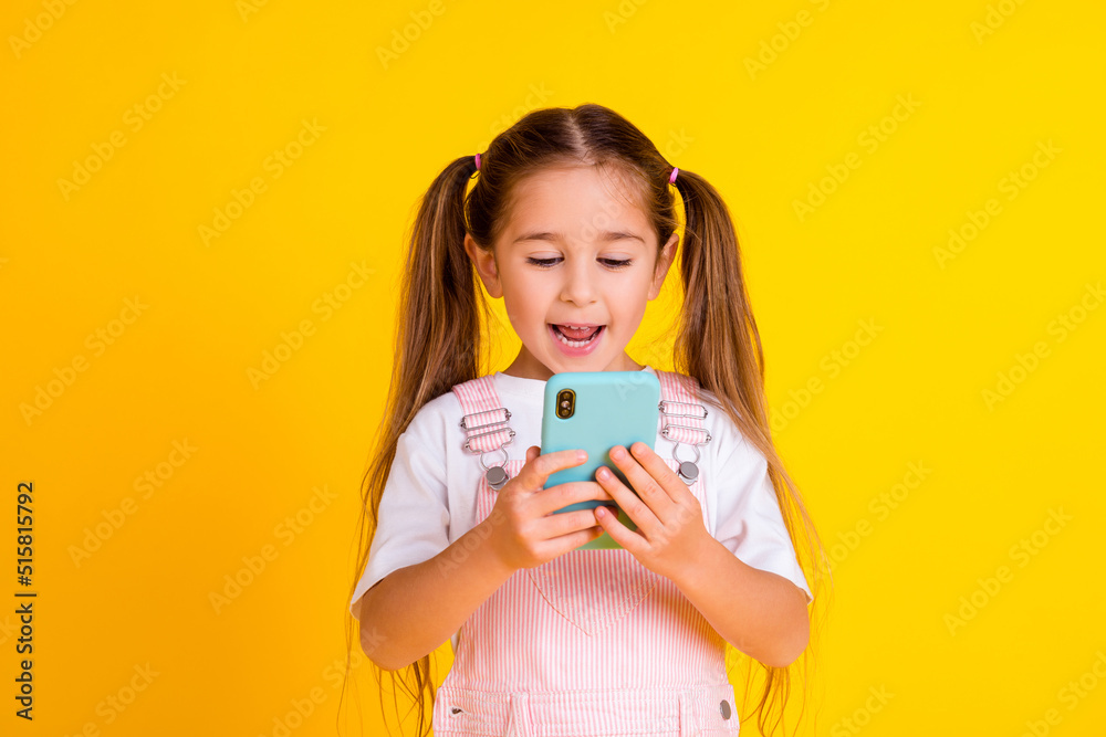 Photo of sweet charming school girl wear summer overall reading modern gadget isolated yellow color background Photo of sweet charming school girl wear summer overall reading modern gadget isolated yellow color background