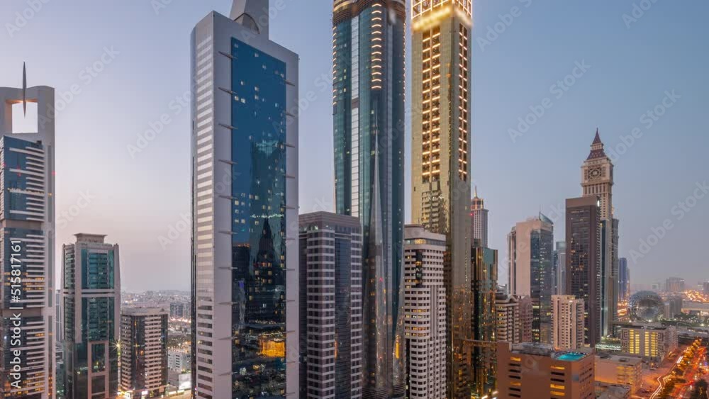 Aerial view of Dubai International Financial District with many skyscrapers day to night transition timelapse after sunset. Traffic on a road near multi storey parking with metro station. Dubai, UAE.