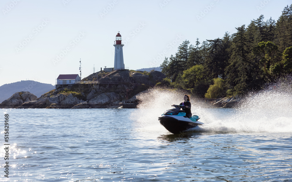 Fototapeta premium Adventurous Caucasian Woman on water scooter riding in the Ocean. Lighthouse park in background. West Vancouver, British Columbia, Canada.