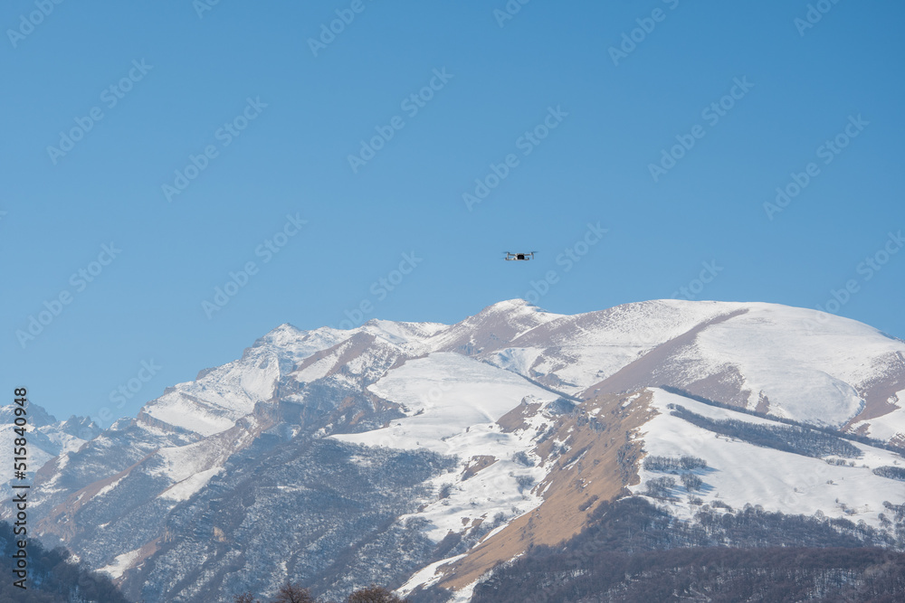 Quadcopter drone flies against the backdrop of snow-capped mountains and blue sky