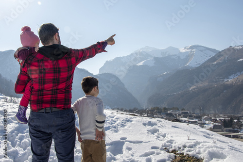 A man in a red plaid shirt stands with children points to the top of the mountain