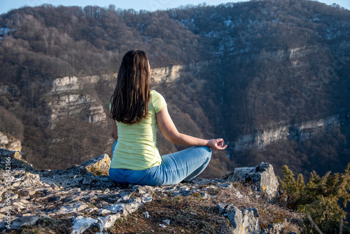 A girl sits on the edge of a cliff in a lotus position, meditates in blue jeans and a yellow T-shirt, view from the back