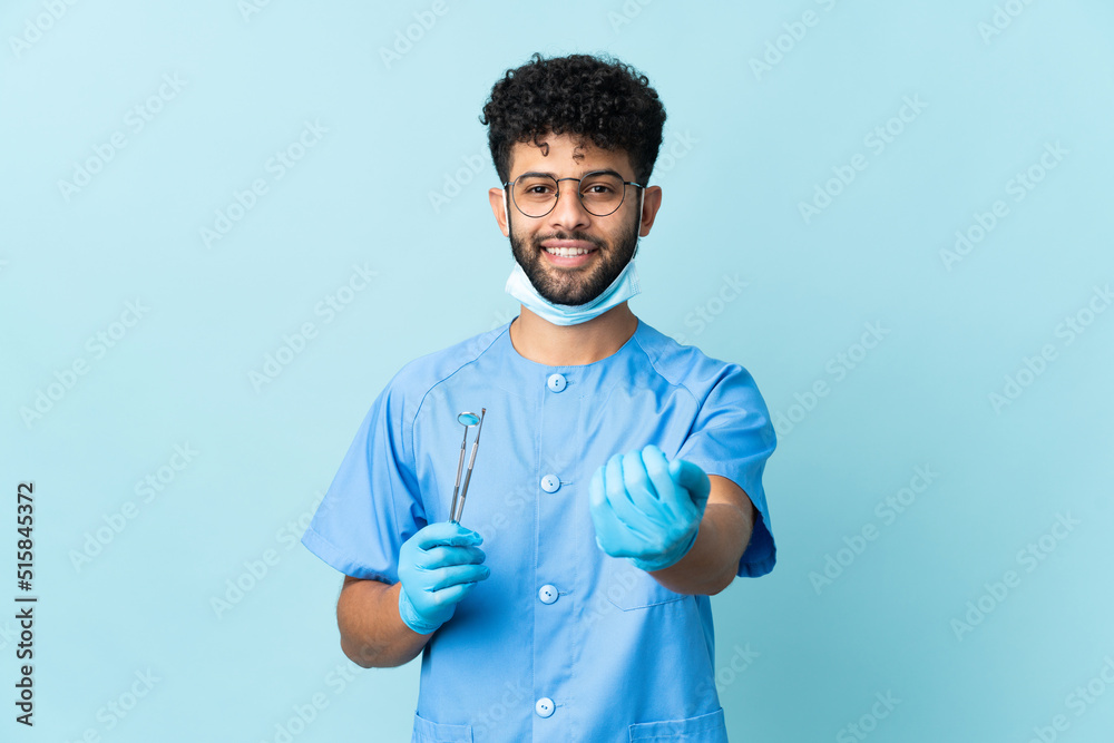 Moroccan dentist man holding tools isolated on blue background inviting to come with hand. Happy that you came Moroccan dentist man holding tools isolated on blue background inviting to come with hand. Happy that you came