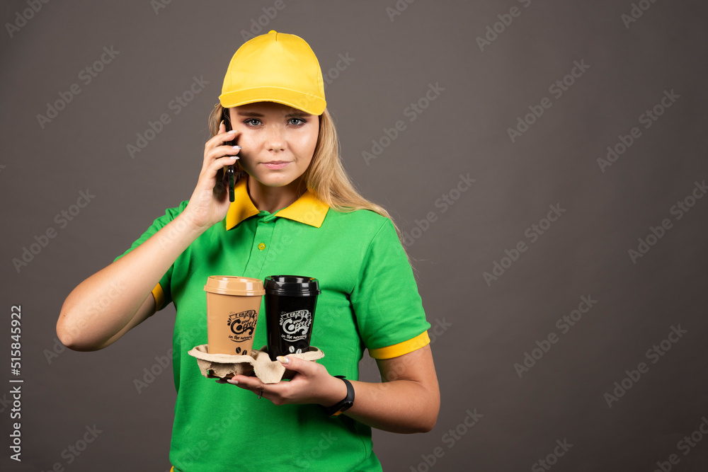 Young deliverywoman with cups of coffee speaking on smartphone