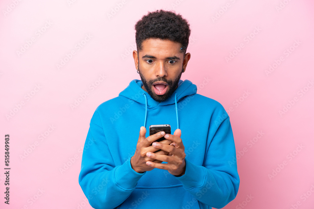 Young Brazilian man isolated on pink background looking at the camera while using the mobile with surprised expression Young Brazilian man isolated on pink background looking at the camera while using the mobile with surprised expression