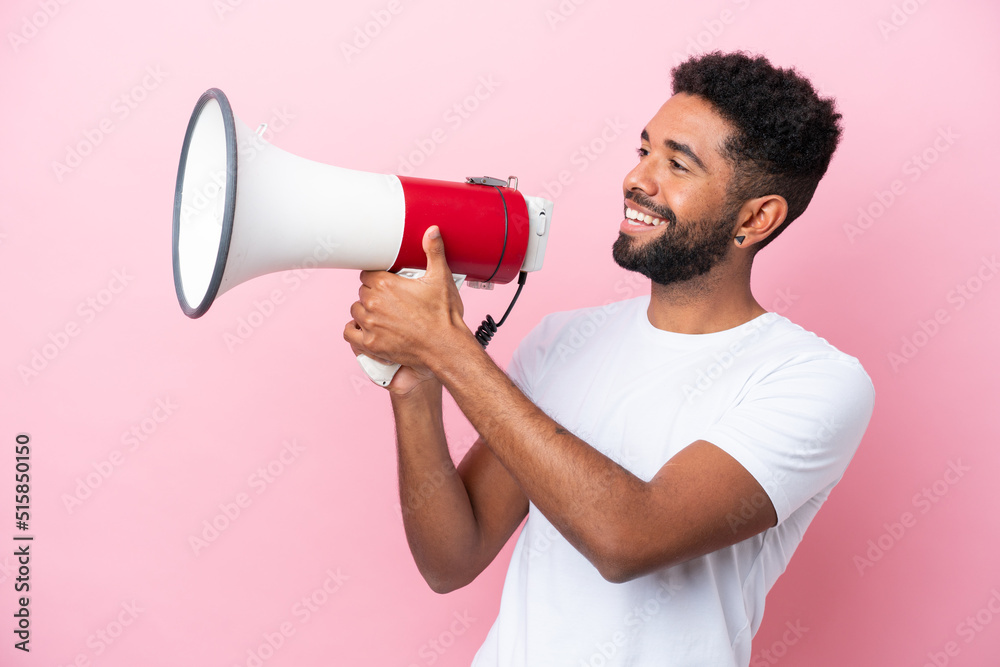 Young Brazilian man isolated on pink background shouting through a megaphone to announce something Young Brazilian man isolated on pink background shouting through a megaphone to announce something