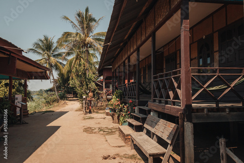 Wooden houses on stilts on a street on Don Det Island, Laos with palm trees in the background on a sunny day.