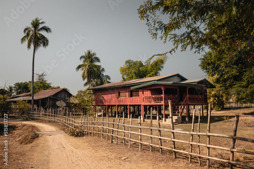 Don Det, Laos - January 18th, 2020 : Colorful red and green lao wooden house on stilts with palm trees in the background and a bamboo fence and sandy pathway in the foreground.