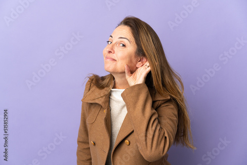 Middle age Brazilian woman isolated on purple background listening to something by putting hand on the ear