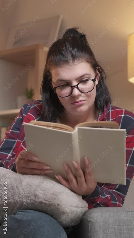 Vertical video. Reading leisure. Home rest. Cozy weekend. Relaxed smart woman in glasses enjoying studying with open book on couch in evening living room. Vertical video. Reading leisure. Home rest. Cozy weekend. Relaxed smart woman in glasses enjoying studying with open book on couch in evening living room.