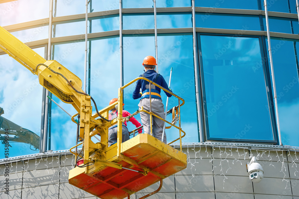 Window washing on high-rise office building in crane bucket. Female team washing glasses windows at height in lifting platform. Window cleaner, building exterior wet wash. Clean service team