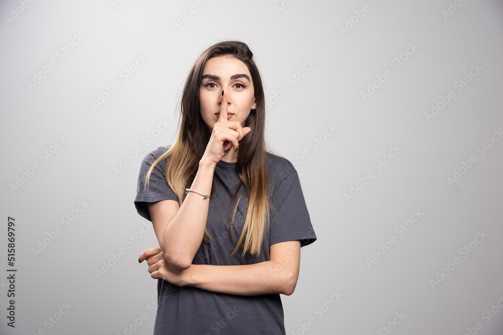 Young woman over a gray background showing a sign of silence gesture putting finger in mouth Young woman over a gray background showing a sign of silence gesture putting finger in mouth