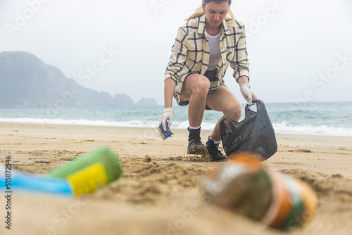 A young woman collects discarded garbage from the beach into a black bag. plastic environmental pollution