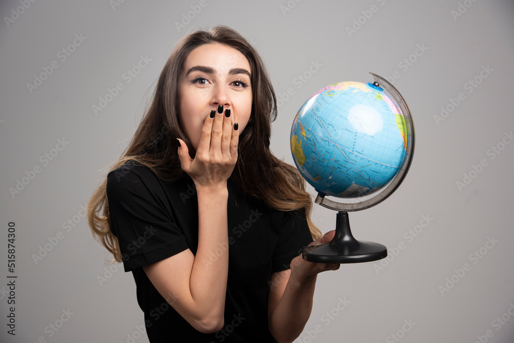 Young woman holding globe on gray background while covering her mouth Young woman holding globe on gray background while covering her mouth