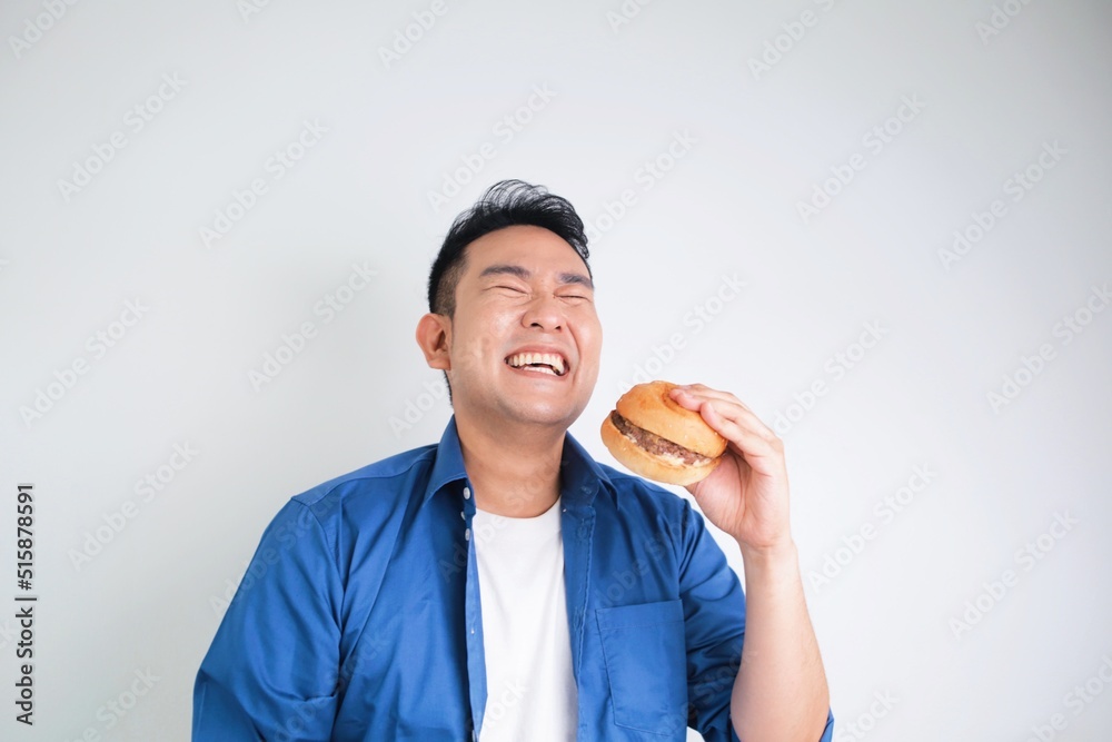 Attractive Asian man in casual shirt holding beef burger standing over white background, copy space. Happy man with burger. Attractive Asian man in casual shirt holding beef burger standing over white background, copy space. Happy man with burger.