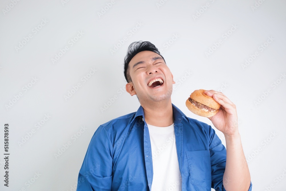 Attractive Asian man in casual shirt holding beef burger standing over white background, copy space. Happy man with burger. Attractive Asian man in casual shirt holding beef burger standing over white background, copy space. Happy man with burger.