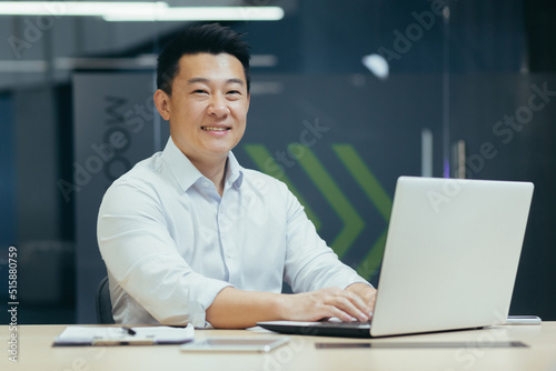 Successful asian businessman working in modern office with laptop typing, smiling and looking at camera.