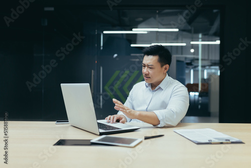 Unhappy and disappointed asian businessman looking at laptop screen and waving hands nervously, man lost money, working in office with laptop.