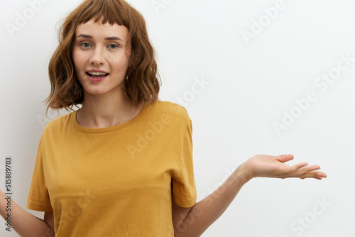 a beautiful, happy, tanned woman with red hair stands on a light background in a yellow t-shirt and looks at the camera making a funny face and spreading her arms to the sides