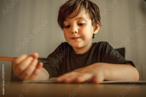 A five - year - old boy is doing his homework . A boy with a pencil in his hands writes, a boy draws on white paper at the table. Primary school and home schooling, the concept of distance education.