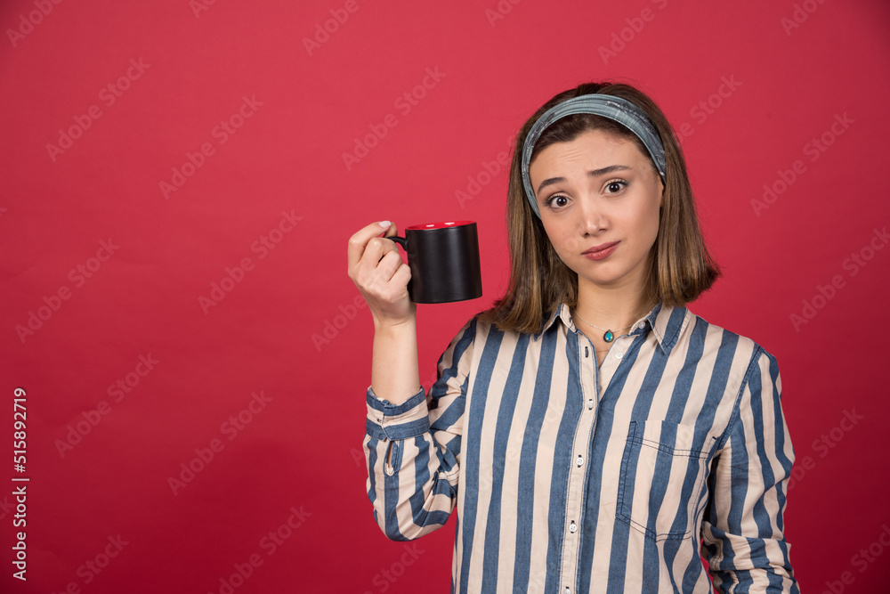 Tired young woman showing cup of coffee and posing to camera Tired young woman showing cup of coffee and posing to camera