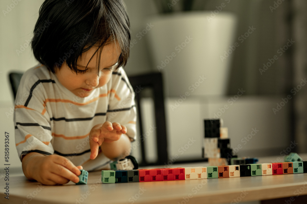 a small Asian boy with long black hair is playing a construction kit while sitting at home at the table. The boy builds from blocks. Educational toys for home and kindergarten. a small Asian boy with long black hair is playing a construction kit while sitting at home at the table. The boy builds from blocks. Educational toys for home and kindergarten.