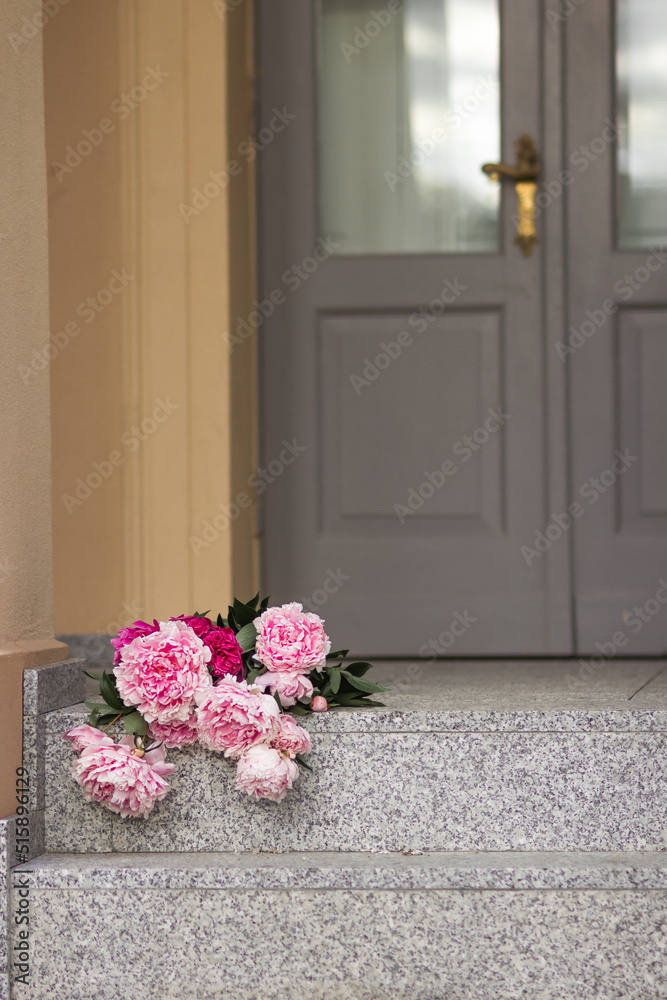 Fototapeta premium Bouquet of pink gardening peonies lies on steps near the old grey wooden door.