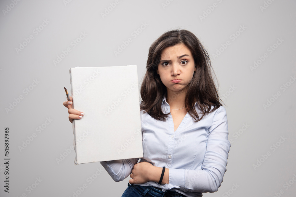 Tired woman showing empty canvas and paintbrush on gray background Tired woman showing empty canvas and paintbrush on gray background