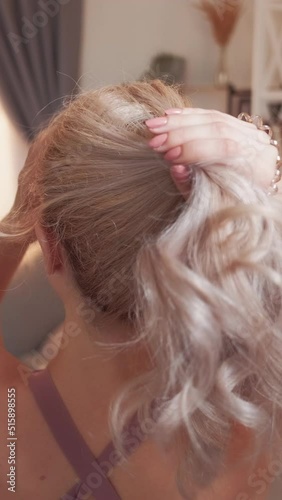 Vertical video. Sporty hairstyle. Blonde woman. Home exercising. Back view of athletic lady tying hair in ponytail light room interior. photo