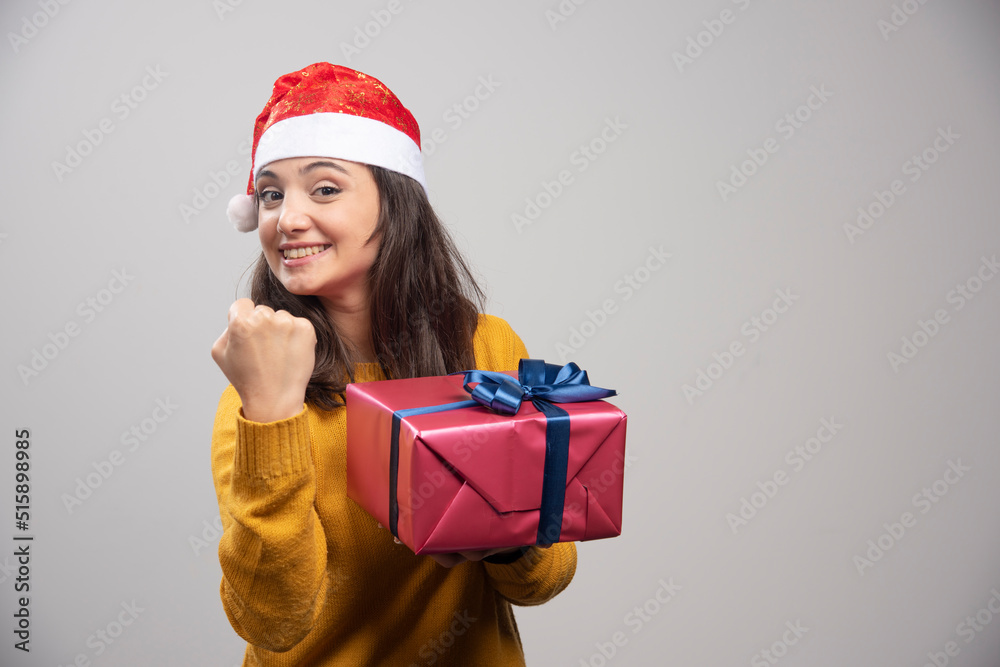 Brunette woman in Santa hat showing her fist and holding gift box Brunette woman in Santa hat showing her fist and holding gift box