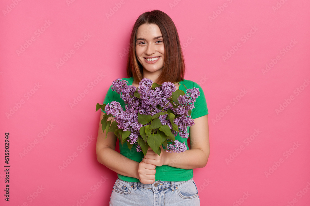 Photo of pleased dark haired woman wearing green T-shirt standing looking with pleasure at camera, carries lilac flowers, models against pink background. Spring present. Photo of pleased dark haired woman wearing green T-shirt standing looking with pleasure at camera, carries lilac flowers, models against pink background. Spring present.