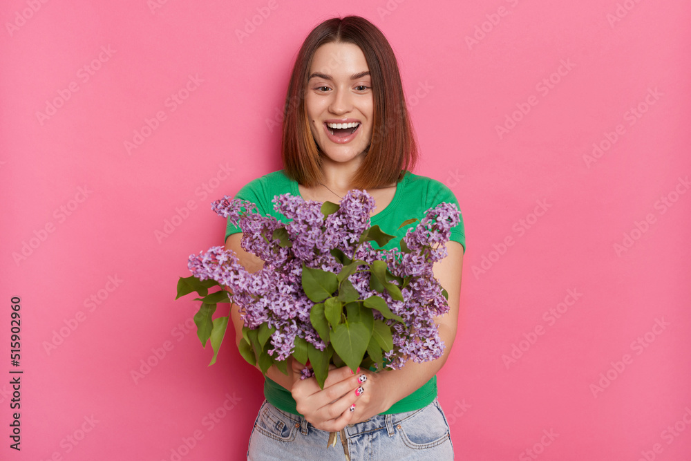 Image of excited woman standing isolated over pink background, looks with amazement at her bouquet, wearing green t shirt, carries lilac flowers, keeps mouth opened, being pleasantly surprised. Image of excited woman standing isolated over pink background, looks with amazement at her bouquet, wearing green t shirt, carries lilac flowers, keeps mouth opened, being pleasantly surprised.